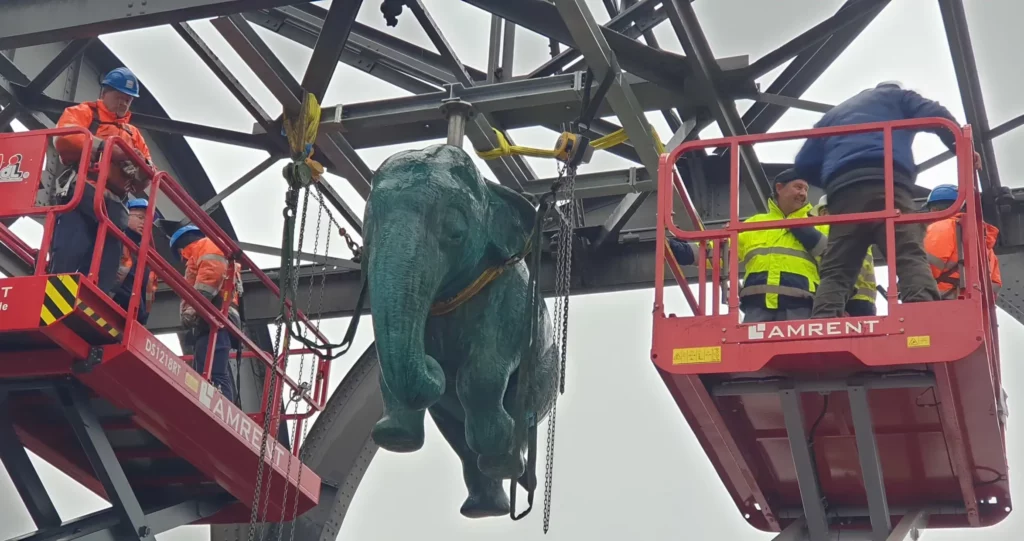 Die Bronzeskulptur eines Elefanten hängt an einem Stahlgerüst zwischen zwei Hebebühnen mit Arbeitern.